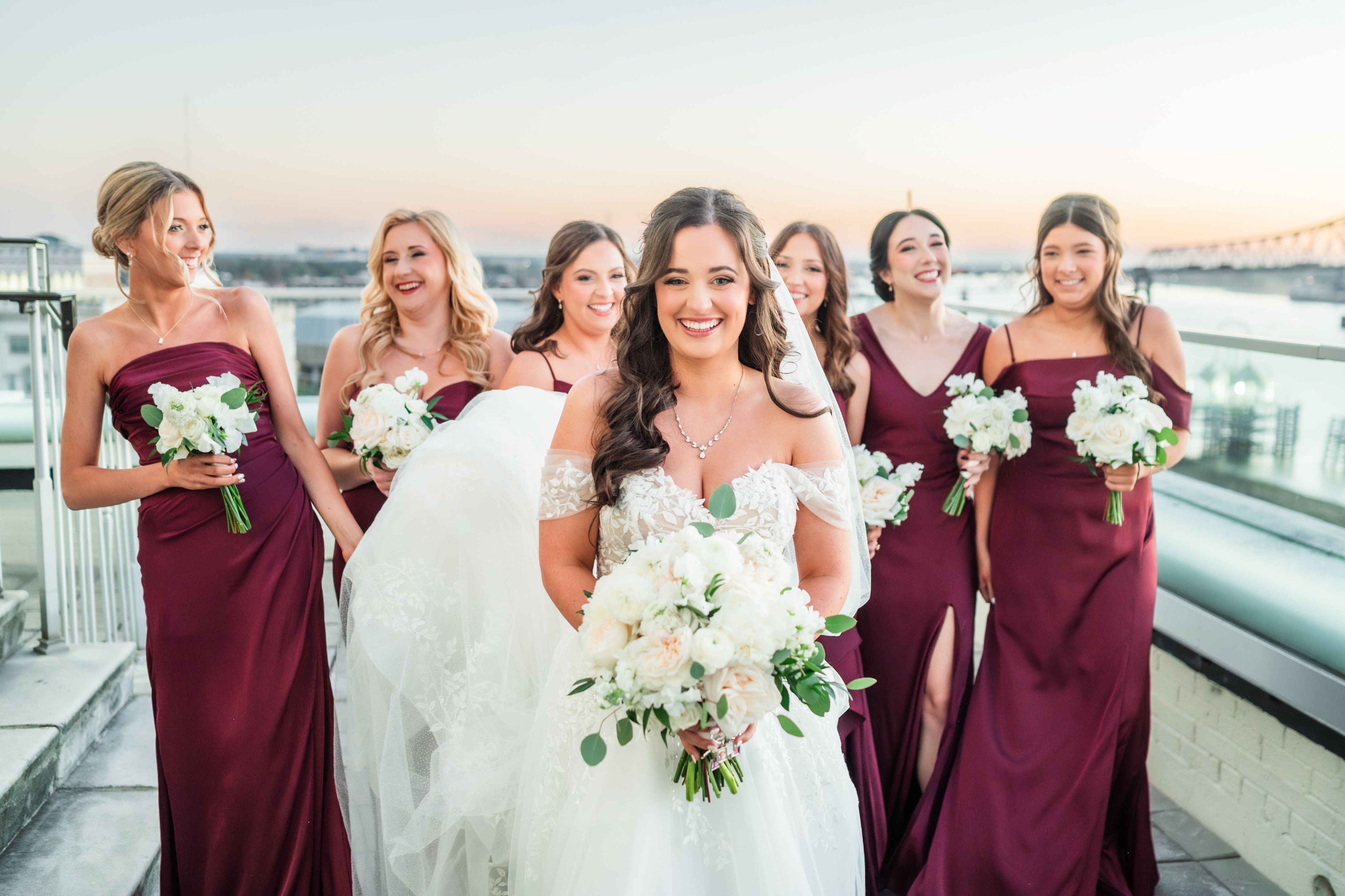 Laughing bride with long waves among bridesmaids in burgundy gowns on a rooftop