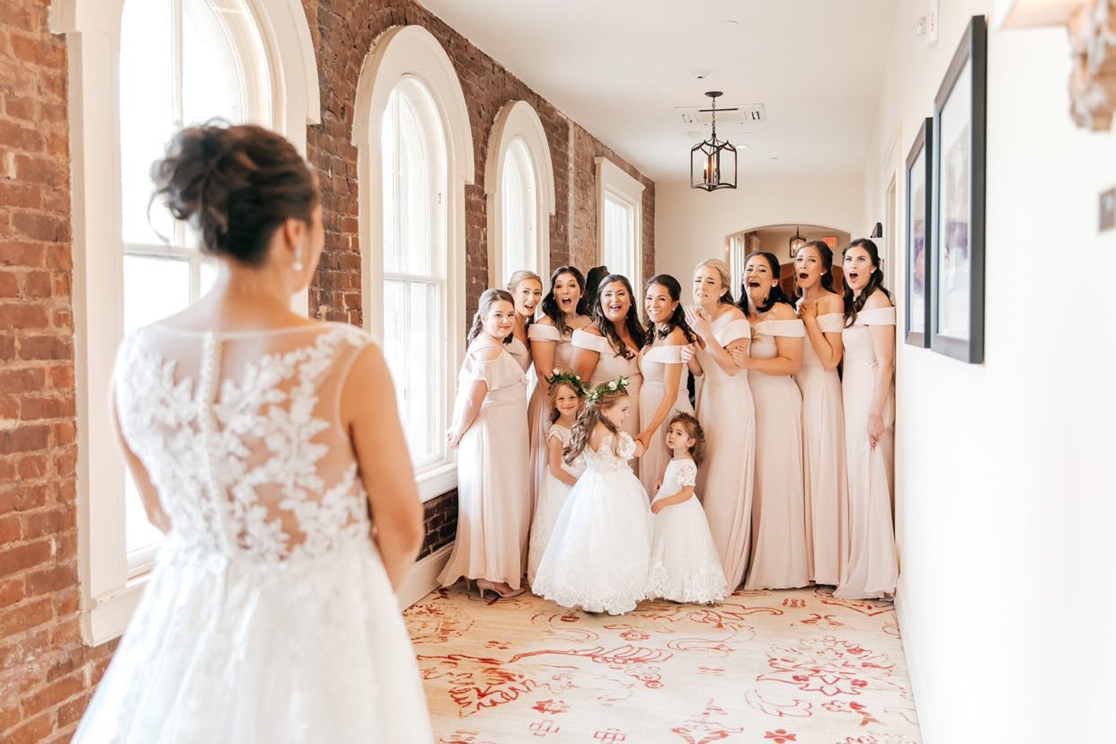 Bridesmaids in blush gowns and flower girls gasping as the bride, in a lace gown with an updo, turns toward them