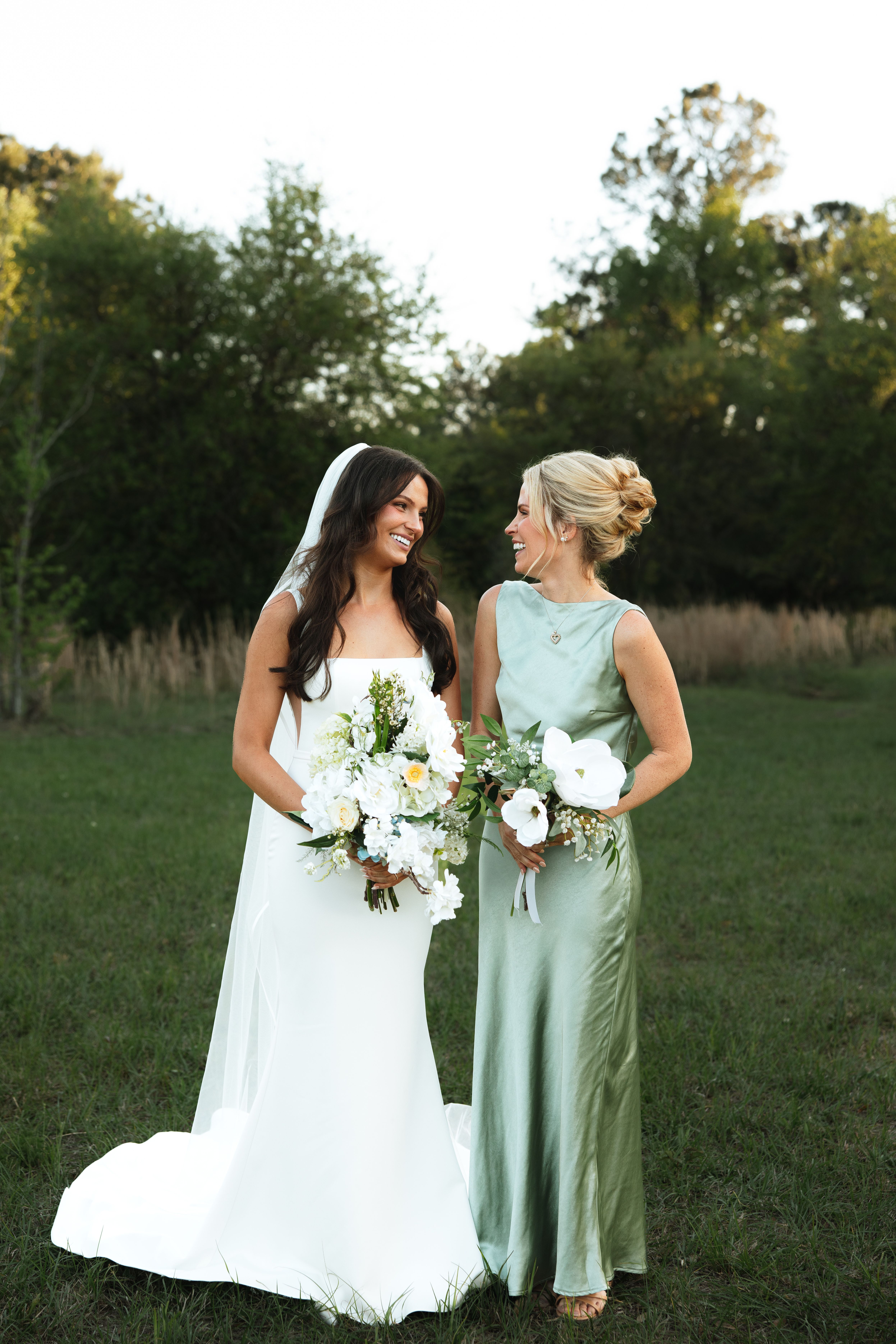Bride with a cathedral veil and long dark waves laughing with a bridesmaid in sage satin with a textured bun