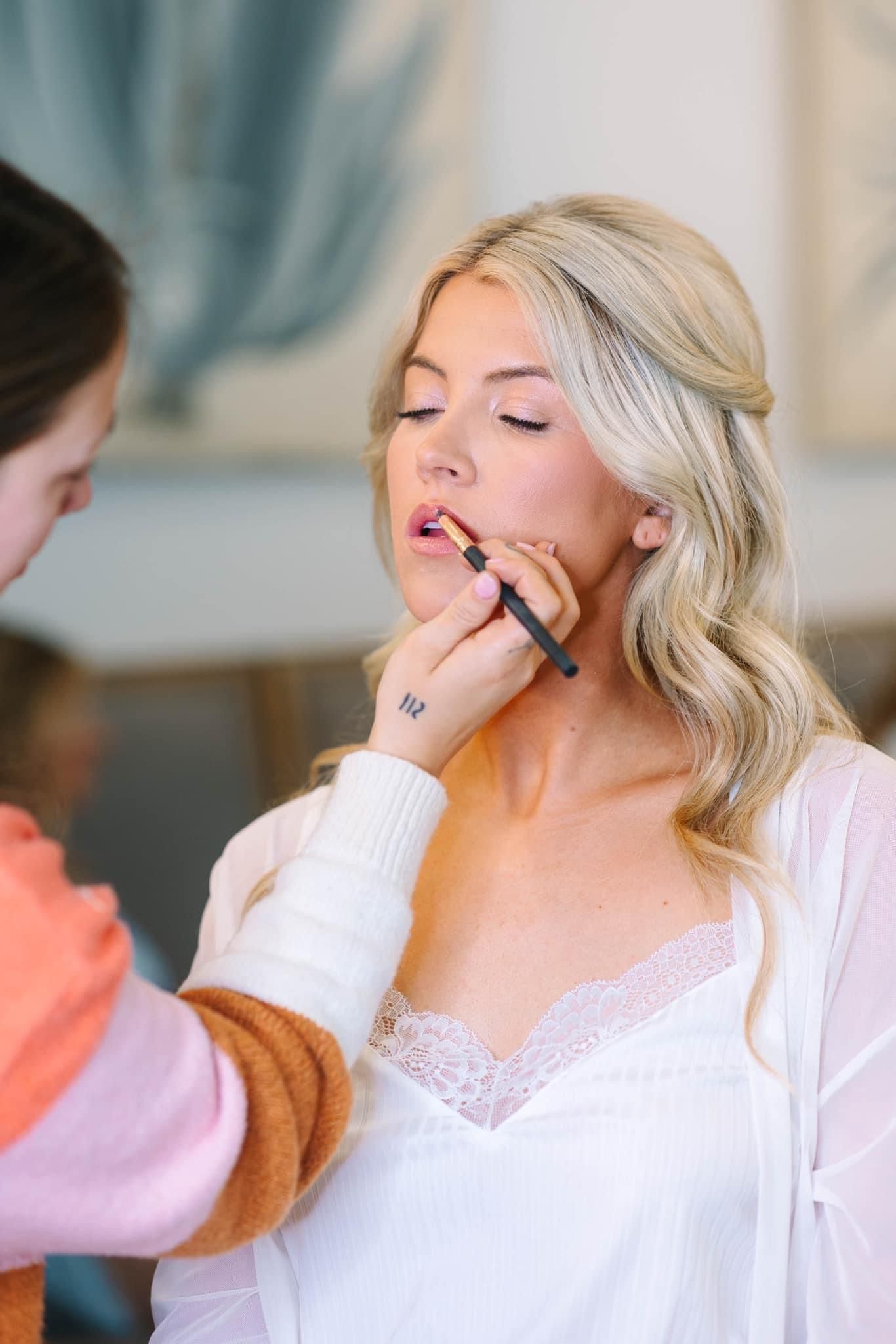 Close-up of lip liner being applied to a bride with soft blonde waves and closed eyes
