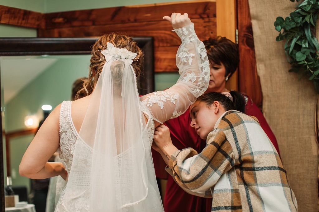 Back of a bride’s curled updo with a floral hair comb and sheer veil as her lace sleeve is fastened