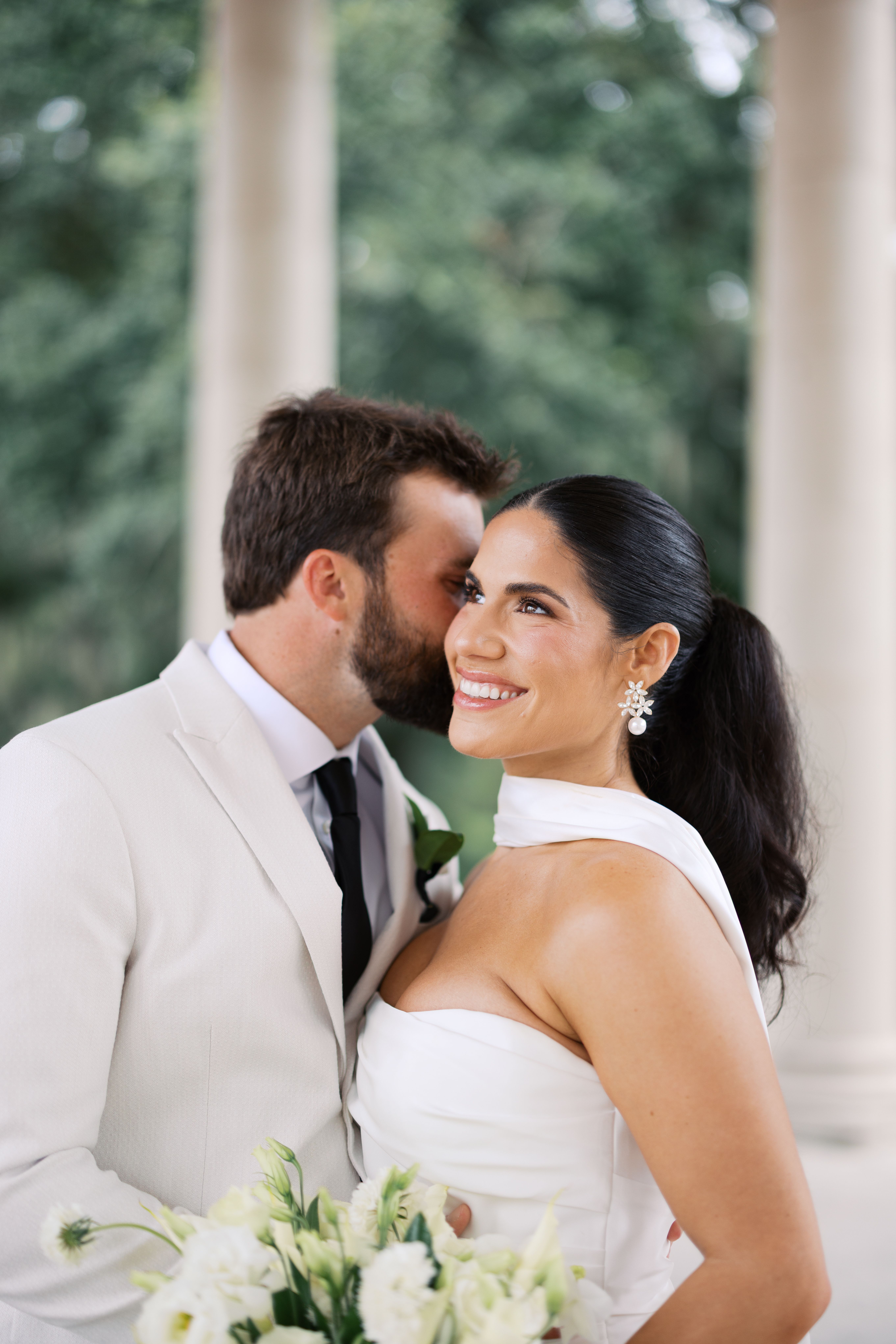 Bride with a sleek low ponytail and pearl earrings smiling as the groom kisses her cheek