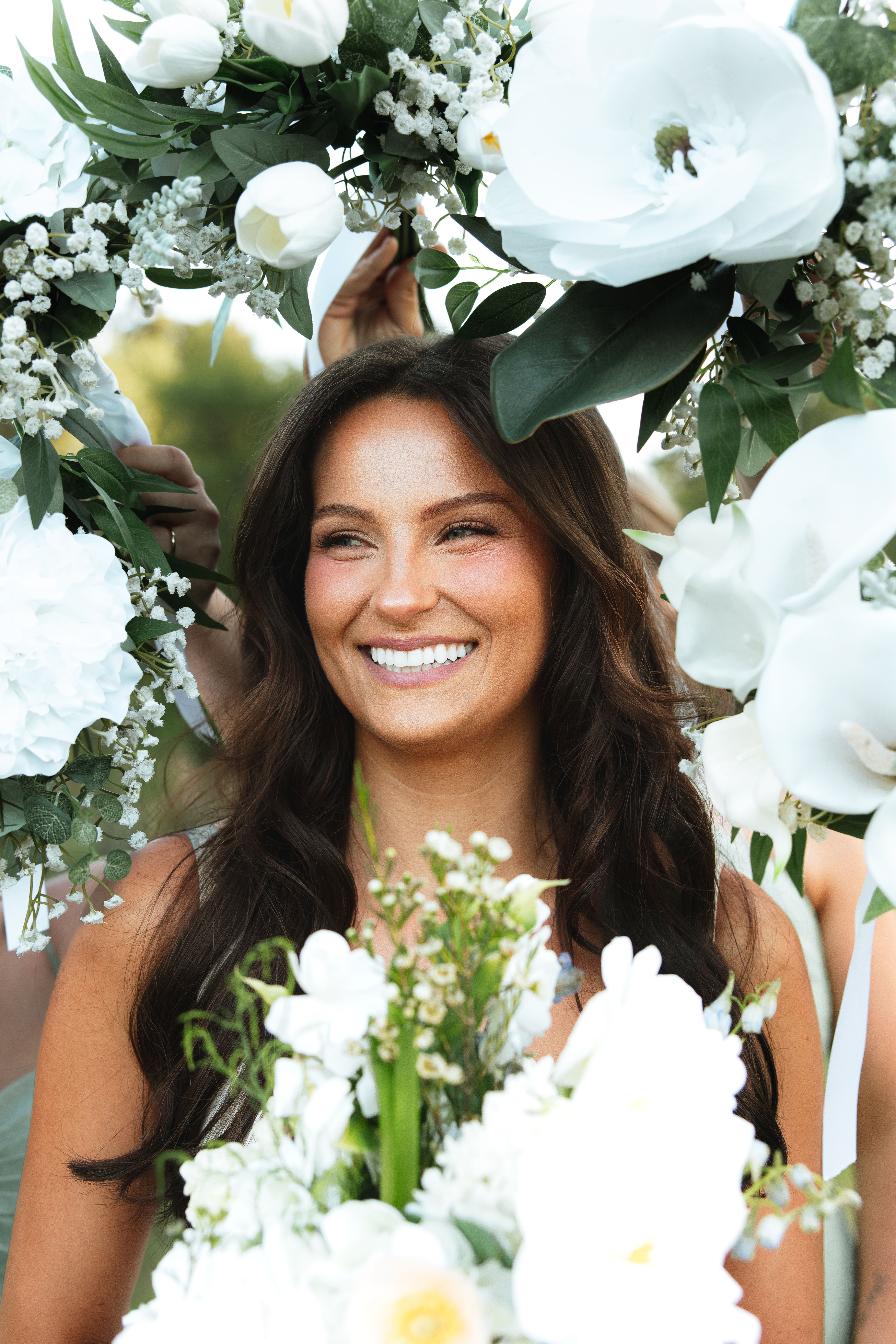 Bride beaming beneath a white floral arch, her long dark hair in loose waves