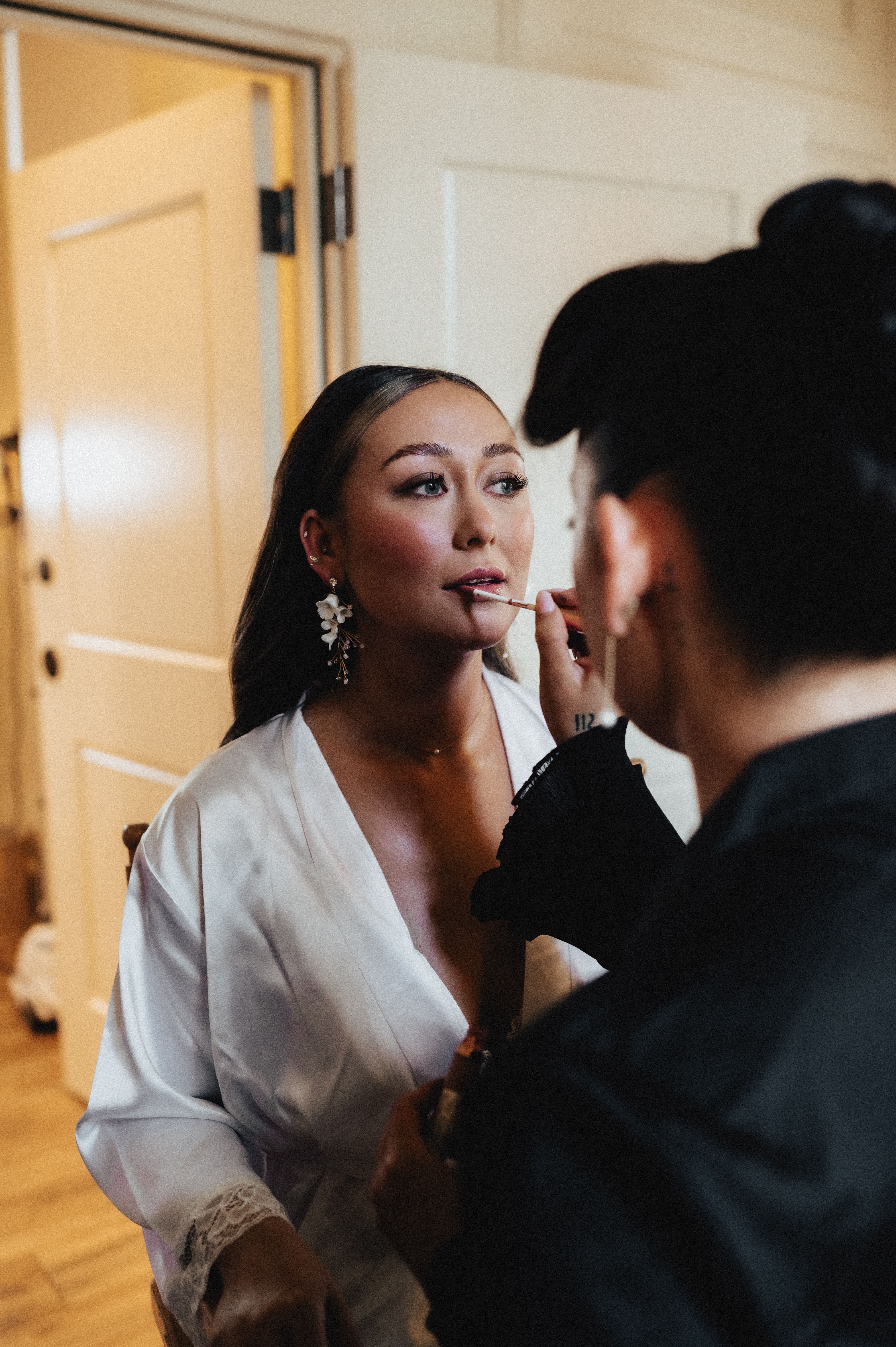 Bride in a satin robe and floral earrings as a makeup artist applies her lip color