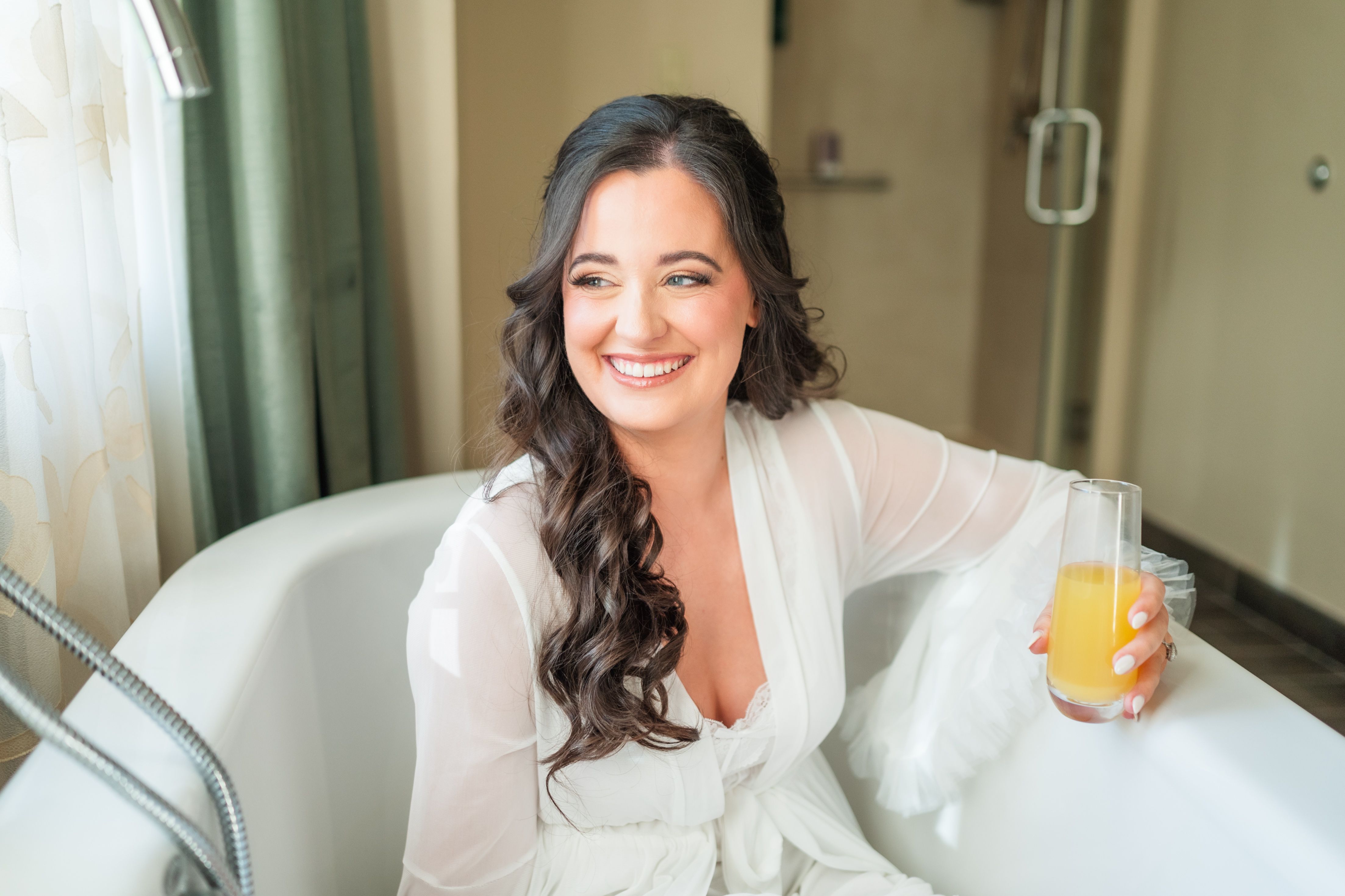 Bride in a white robe with loose brunette curls smiling from a soaking tub, mimosa in hand