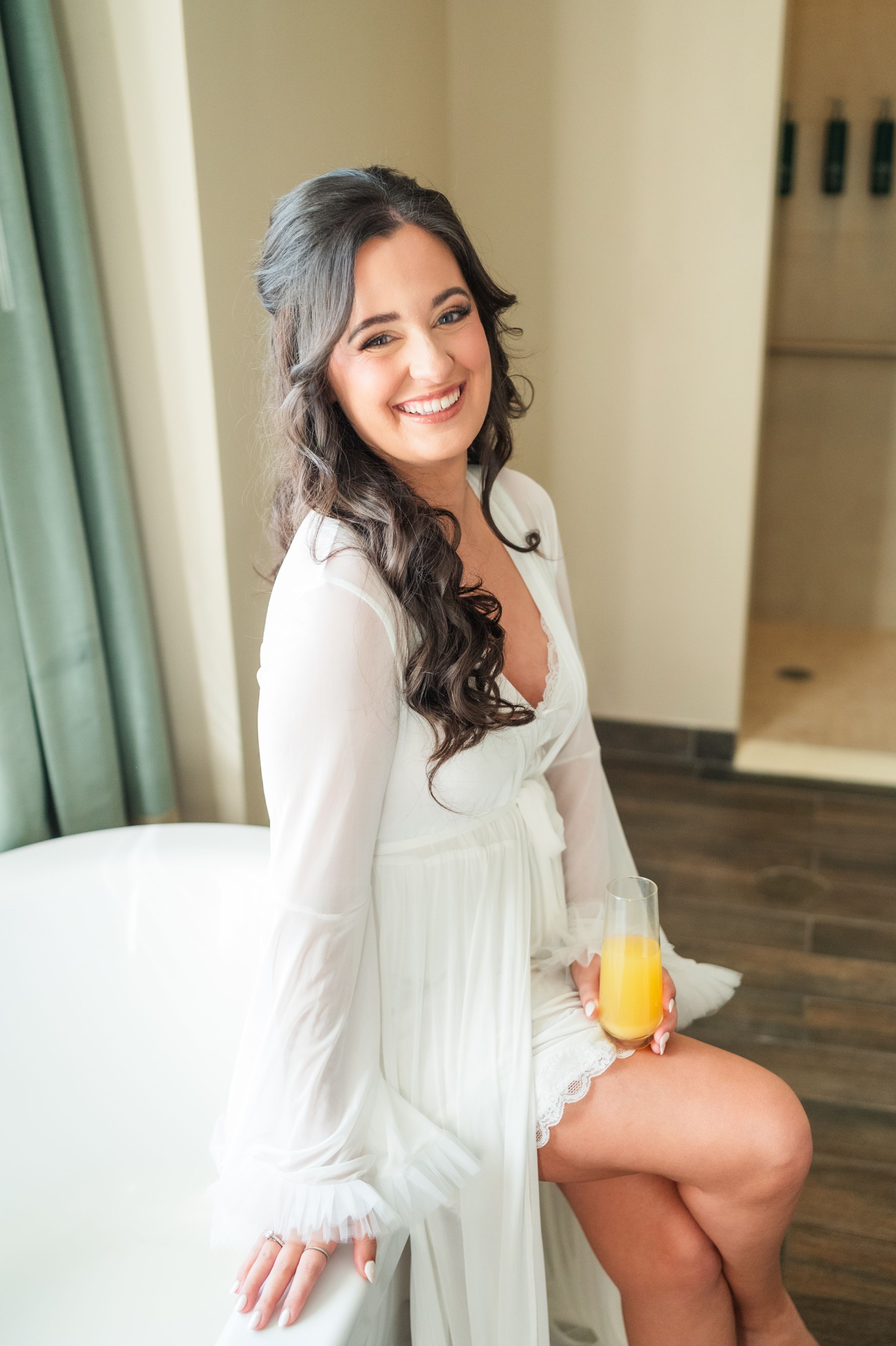 Bride laughing in a sheer white robe with loose brunette curls, holding a mimosa