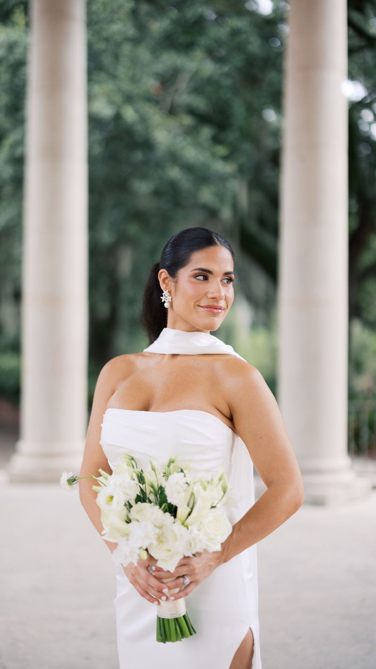 Bride with a sleek low ponytail and a scarf-neck gown holding white flowers among stone columns