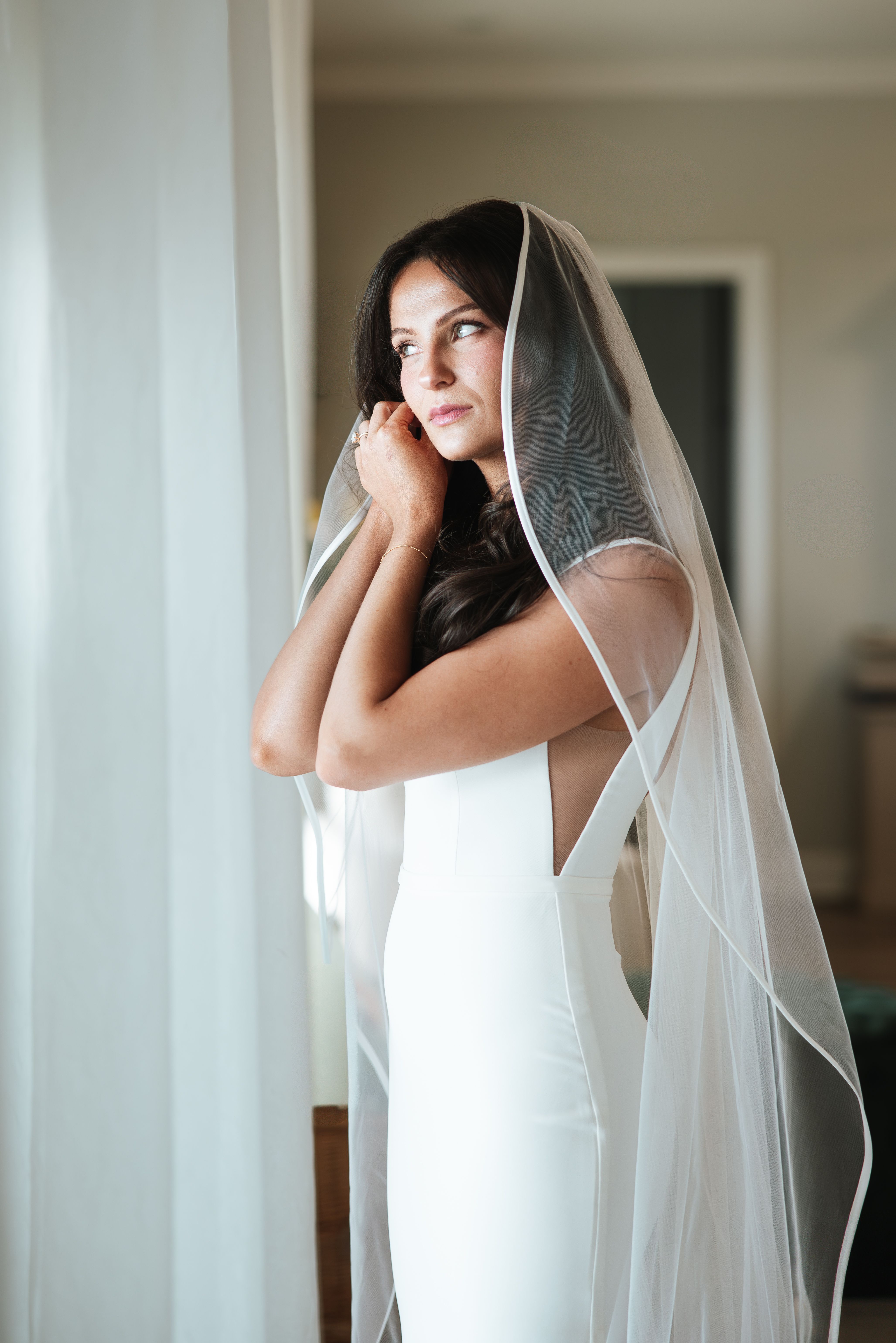Bride with long dark waves and a sheer veil fastening an earring beside a window