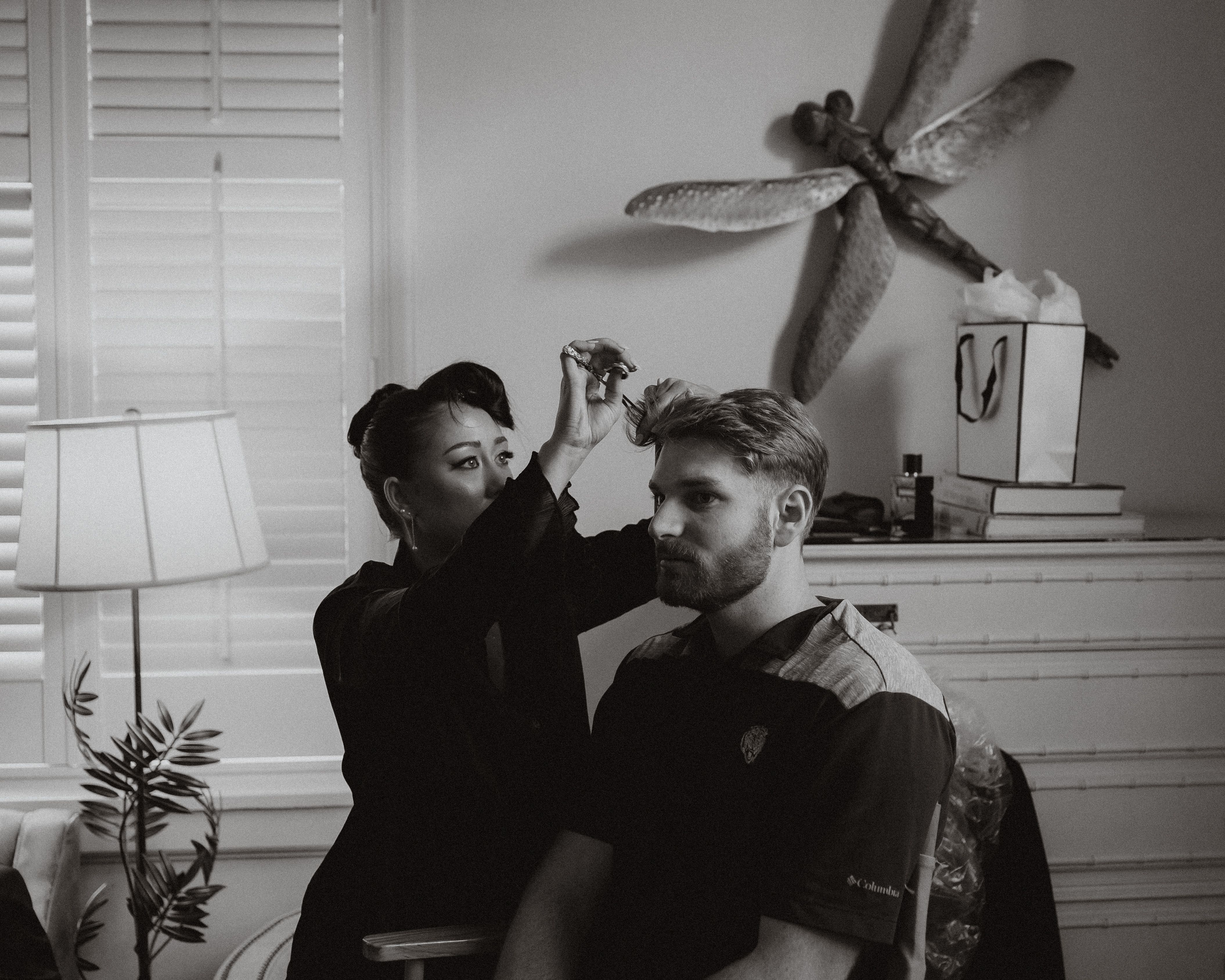Black-and-white photo of a stylist trimming a bearded client’s hair with scissors